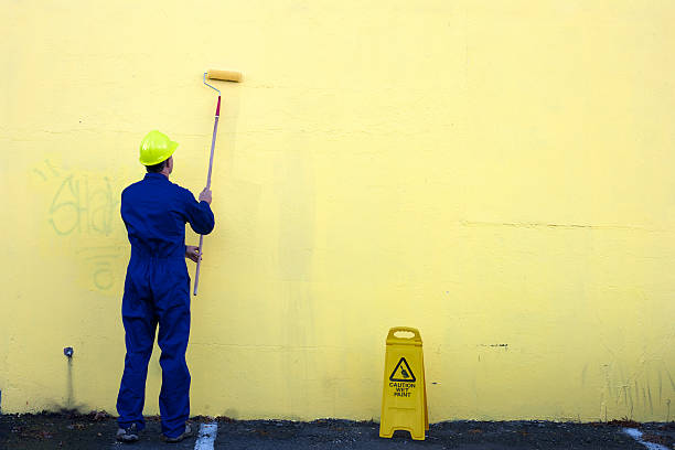 A man paints over graffiti on a yellow wall outdoors.
