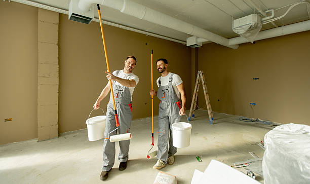Two men in gray overalls are smiling and holding paint rollers and buckets. They are in a bright office with freshly painted walls. The atmosphere is friendly and upbeat.